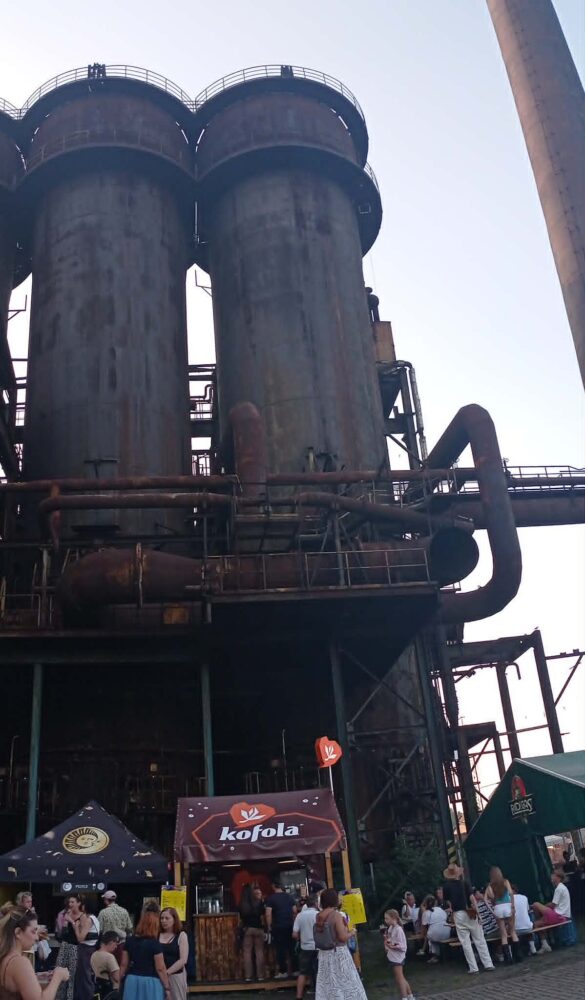 Industrial factory towers with pipes and scaffolding behind a festival crowd under a clear sky.
