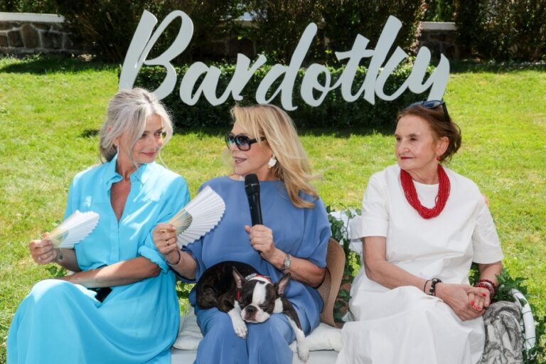 Three women sit outdoors at an event with a large 'Bardotty' sign behind them; the middle woman holds a microphone and a small dog rests on her lap.