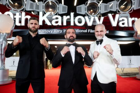 Three men in tuxedos pose with fists raised on a red carpet at the Karlovy Vary International Film Festival.
