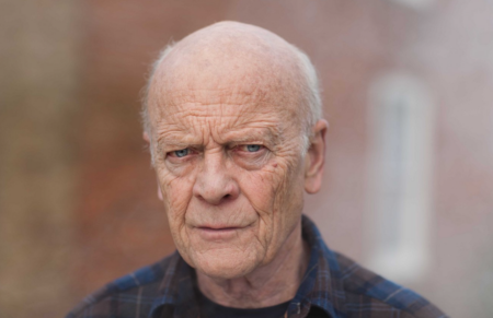 Older man with a stern, thoughtful expression, wearing a blue plaid shirt outdoors.