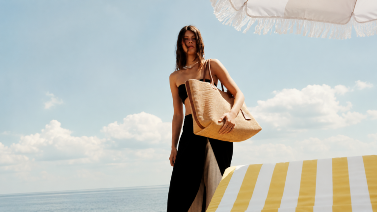 Woman in a black outfit carrying a large woven straw tote bag at the beach, with a yellow-and-white striped umbrella in the foreground.