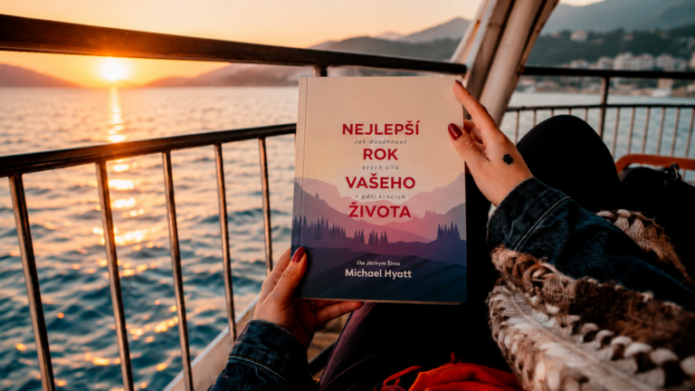 A person in a denim jacket holds a book titled 'Nejlepší rok vašeho života' aboard a boat at sunset over calm water.