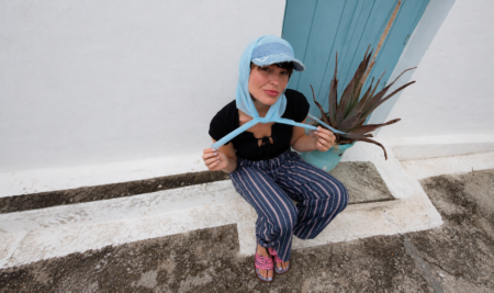 Woman sitting on concrete steps against a light blue wall, wearing a blue scarf, denim cap, black top, striped pants, and pink sandals beside a potted plant.