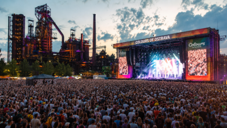 Large crowd at an outdoor concert with a colorful, illuminated stage and an industrial backdrop at dusk.
