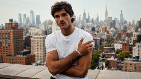 Young man in a white T-shirt standing on a rooftop with a city skyline in the background, gazing at the camera.