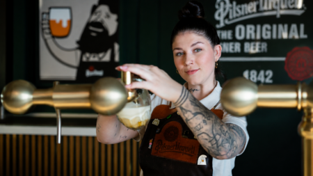 Female bartender with a sleeve tattoo and a Pilsner Urquell apron pours beer from a tap at a bar.