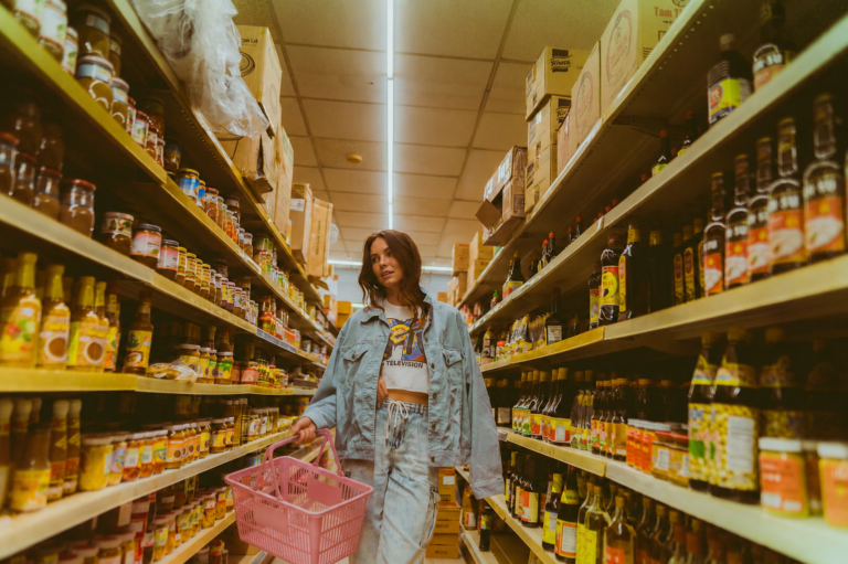 Young woman with a pink shopping basket browses shelves lined with bottles and jars in a grocery store aisle.