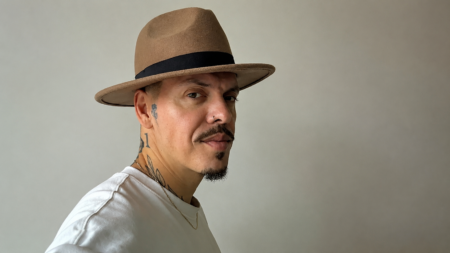 Man wearing a tan fedora with a black band, facing slightly to the right, against a neutral backdrop.
