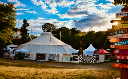 Large white circus-like tent at an outdoor festival, with banners along the fence and a signpost on the right under a blue sky.