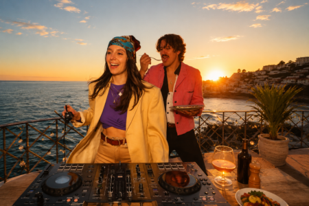 Couple at sunset on a balcony with a DJ mixer, woman in yellow blazer dancing, man feeding her a bite while holding a plate and wine nearby