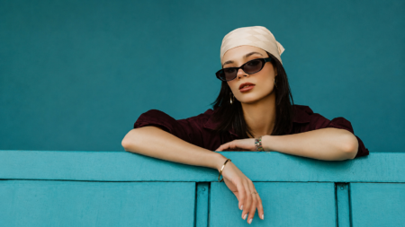 Portrait of a woman in a beige headscarf and black sunglasses leaning on a turquoise railing, wearing a burgundy top and gold jewelry.