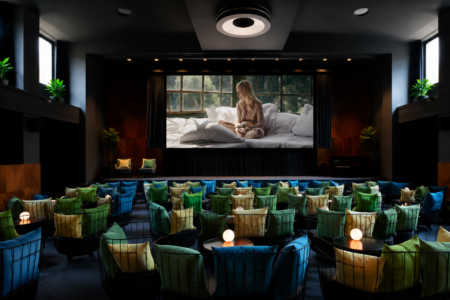 Screening room with a large projection showing a woman sitting on a bed by a window; foreground has rows of colorful cushioned chairs and small round lamps on tables.