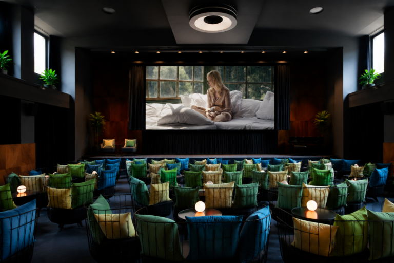 Screening room with a large projection showing a woman sitting on a bed by a window; foreground has rows of colorful cushioned chairs and small round lamps on tables.