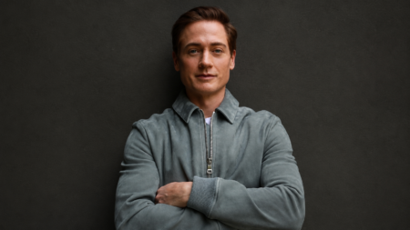 Portrait of a confident man with arms crossed, wearing a gray zippered jacket against a dark wall.