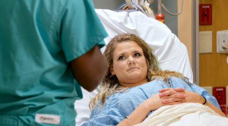 Female patient in a blue hospital gown lying in bed, hands folded, talking with a healthcare professional in teal scrubs beside the bed.