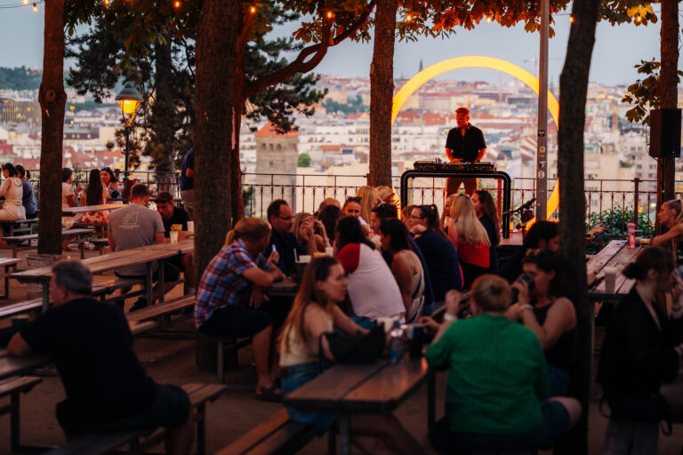 Crowd gathered at picnic tables on a rooftop at dusk while a DJ plays near a bright circular orange light and city skyline.