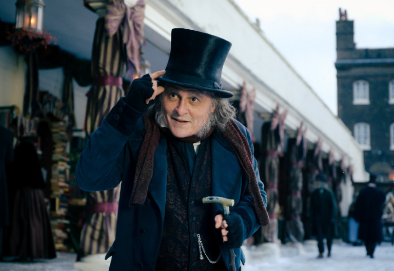 Man in a dark top hat tipping it in a Victorian-era coat at a snowy outdoor market, holding a cane.