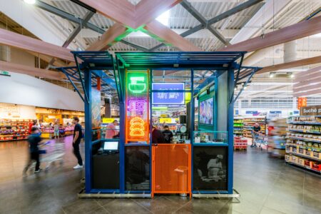 Neon-lit display kiosk in a modern supermarket center with shoppers and aislesAround a blue and orange metal structure under wooden beams.