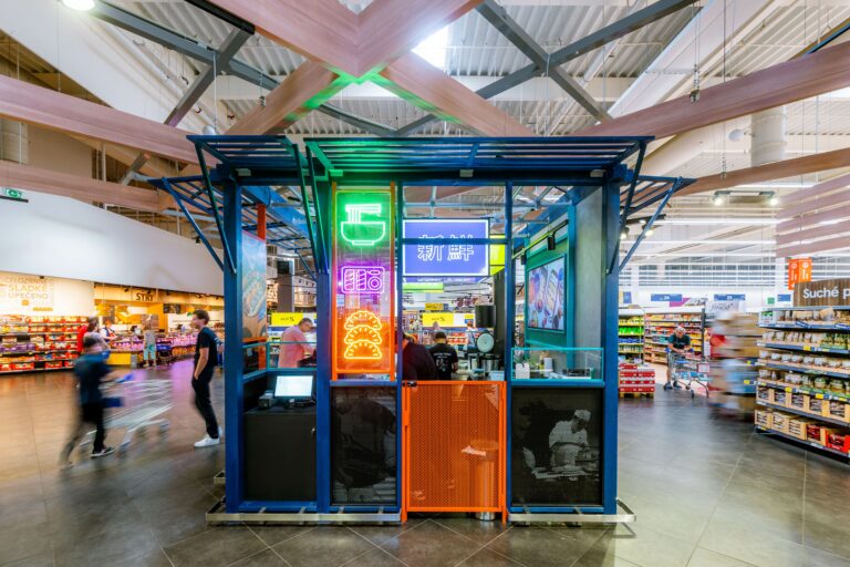 Neon-lit display kiosk in a modern supermarket center with shoppers and aislesAround a blue and orange metal structure under wooden beams.