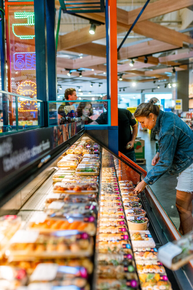 Woman in a denim jacket leaning over a long refrigerated display of pre-packaged sushi and bowls in a grocery store; other shoppers in the background; neon signs on left side.
