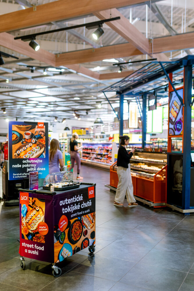 Colorful street-food sushi cart in a supermarket aisle, with displays of sushi and shoppers in the background.