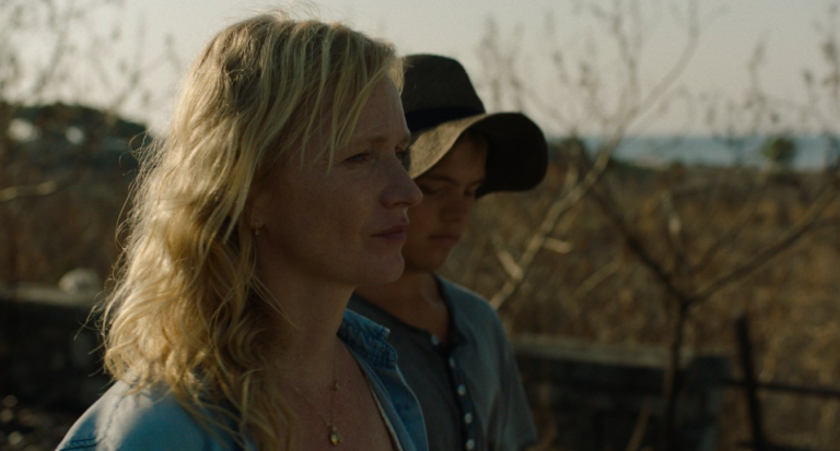 A woman with wavy blonde hair in the foreground, standing outdoors with a man wearing a hat behind her.