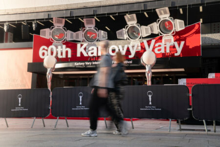 Red banner reading '60th Karlovy Vary' at the festival entrance with barricades and spotlights above; people blurred as they walk by.