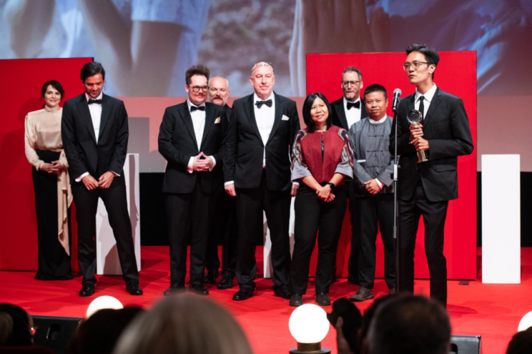 Group of formally dressed people on a red-carpet stage during an award ceremony; a man on the right holds a trophy and speaks into a microphone.