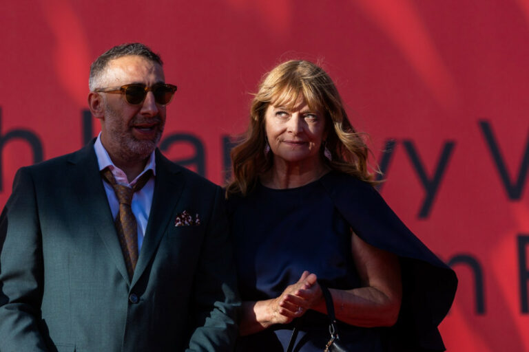 Man in teal suit and brown tie with sunglasses stands beside a woman in a navy dress at a red backdrop event.