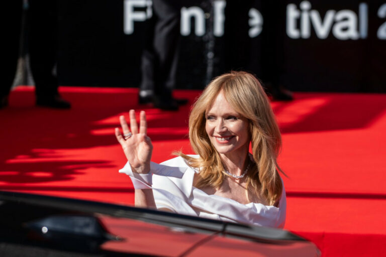 Woman in a white off‑the‑shoulder dress smiles and waves from a red carpet event.
