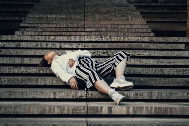 Person lying on concrete stairs, wearing a white shirt, black-and-white striped skirt, and sneakers.