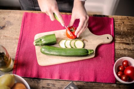 Person slicing a tomato on a wooden cutting board with zucchini beside it on a red placemat.