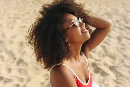 Woman with curly hair wearing sunglasses and a red swimsuit, smiling at the beach.