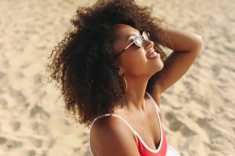 Woman with curly hair wearing sunglasses and a red swimsuit, smiling at the beach.
