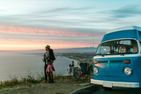 Couple standing beside a bright blue vintage van at sunset overlooking the ocean.
