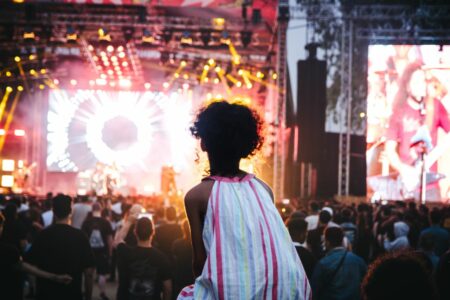 Back view of a person with curly hair in a pastel-striped garment standing among concertgoers facing a bright stage.
