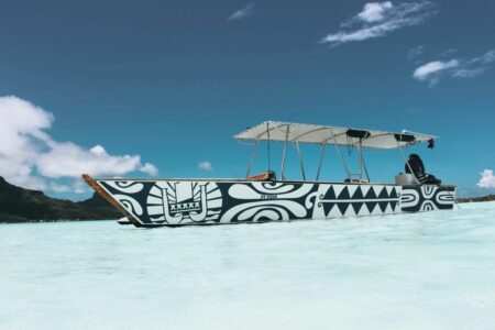 A long white boat with bold black tribal patterns floats on crystal turquoise water with a clear blue sky above, a person seated toward the rear under a canopy.