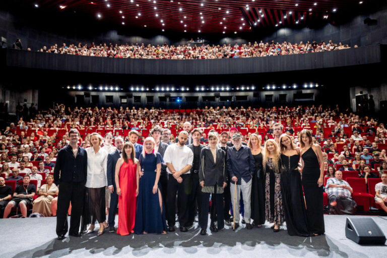 Group of performers standing on a theater stage for a photo with a large audience in red seats behind them.