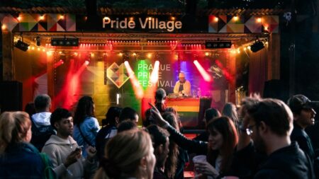 DJ performing on a stage at a Pride Village festival, surrounded by colorful lights and a cheering crowd at dusk/night.