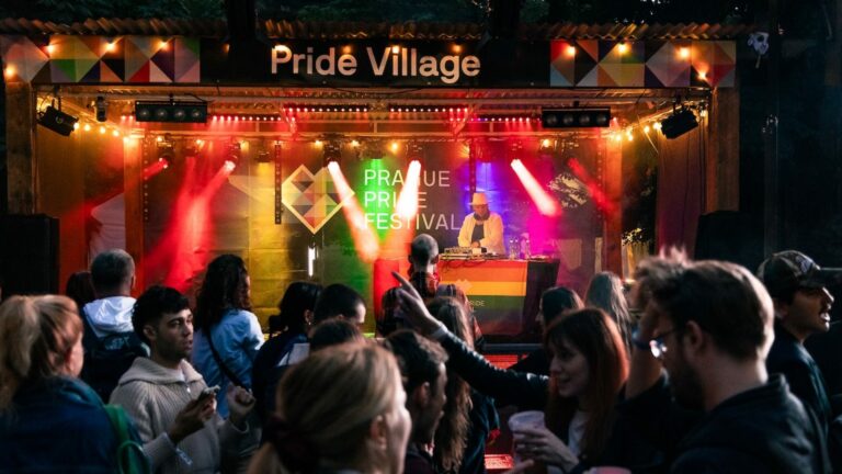 DJ performing on a stage at a Pride Village festival, surrounded by colorful lights and a cheering crowd at dusk/night.