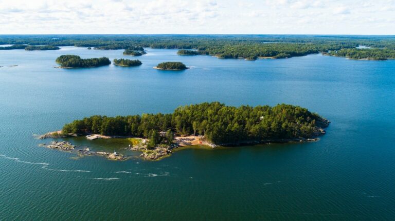 Aerial view of a forested island with rocky shores in a calm blue lake, with more small islands in the distance under a partly cloudy sky.