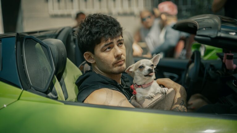 Man with short dark hair riding in a lime-green convertible, holding a small white dog with a pink collar.