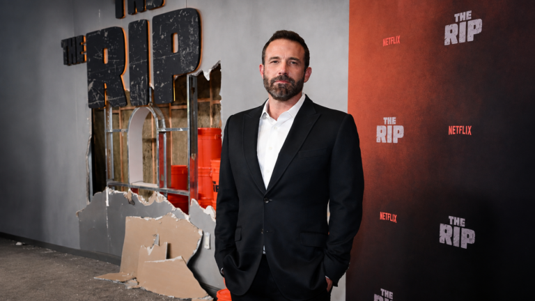 Man in a dark suit and white shirt posing on a red-carpet backdrop for The Rip Netflix event, with a damaged wall set to the left.
