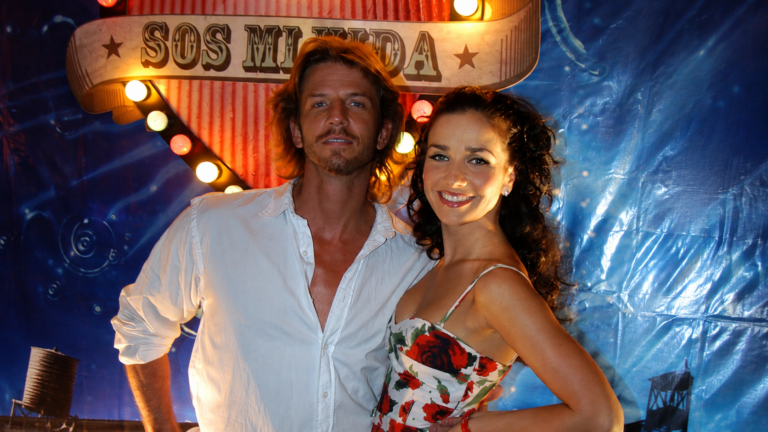 Man and woman posing together at a carnival-style backdrop with lights and a banner above them.