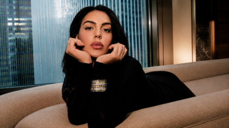 Woman with long dark hair in a black top, resting her chin on her hands as she lounges on a beige sofa in a modern, city-view room.