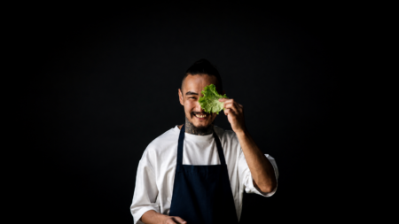 Chef wearing a white shirt and navy apron holds a leafy head of lettuce near his eye, smiling against a dark background.