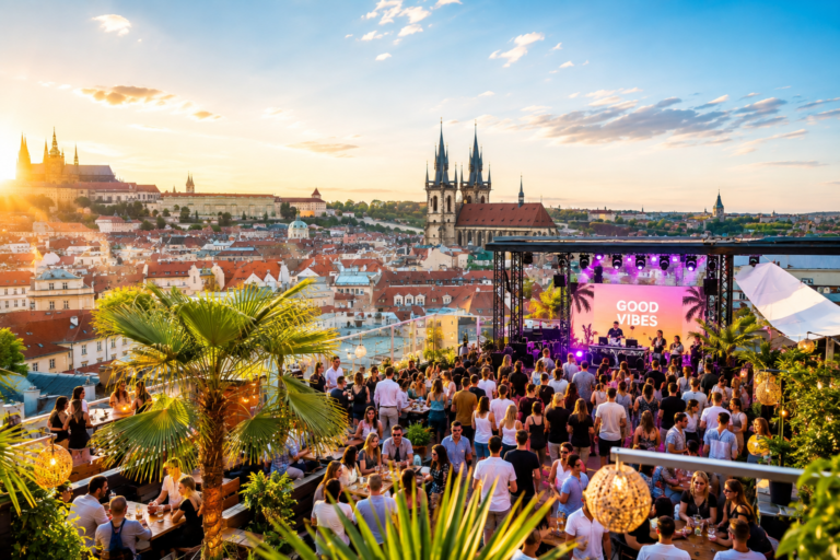 Crowd on a rooftop terrace at sunset watching a live band on a stage with a 'GOOD VIBES' screen, overlooking a historic European city.