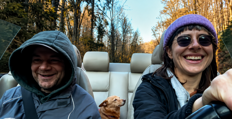 Two people riding in a cream-colored convertible with a small tan dog between them, smiling as they drive through a forest road.