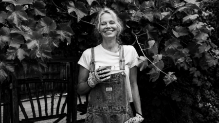 Smiling woman in denim overalls holding a mug, standing among leafy grapevine leaves.