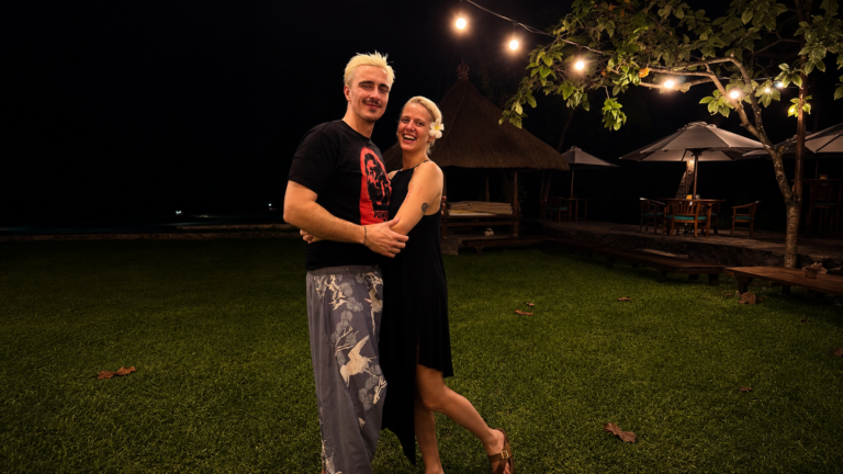 Couple dancing and embracing on a grassy lawn at night, string lights overhead and a tropical seating area in the background.